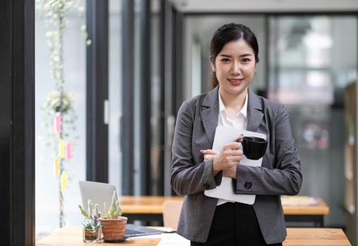 Charming Asian woman with a smile standing holding tablet and cup at the office..