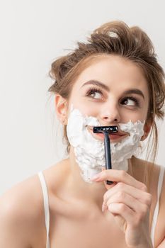 Beautiful young caucasian smiling woman shaving her face with razor looking up on white background