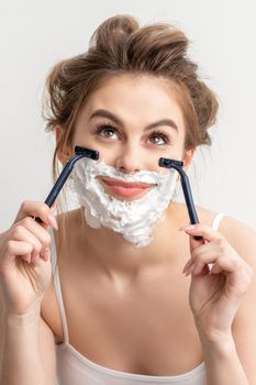 Beautiful young caucasian smiling woman shaving her face with razor looking up on white background