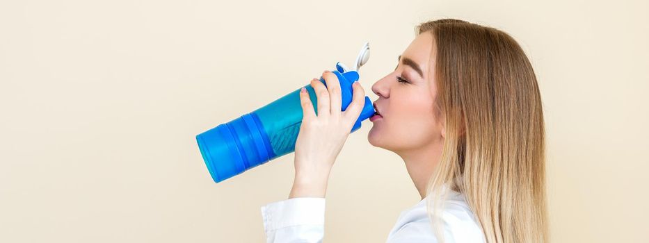 Side view of beautiful young caucasian woman is drinking water from plastic bottle against a beige background