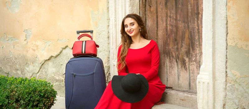 A young caucasian woman is sitting on the threshold of an old house in a red long dress with suitcase looking at the camera outdoors