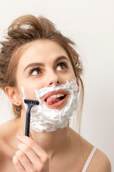 Beautiful young caucasian smiling woman shaving her face with razor sticking out tongue looking up on white background