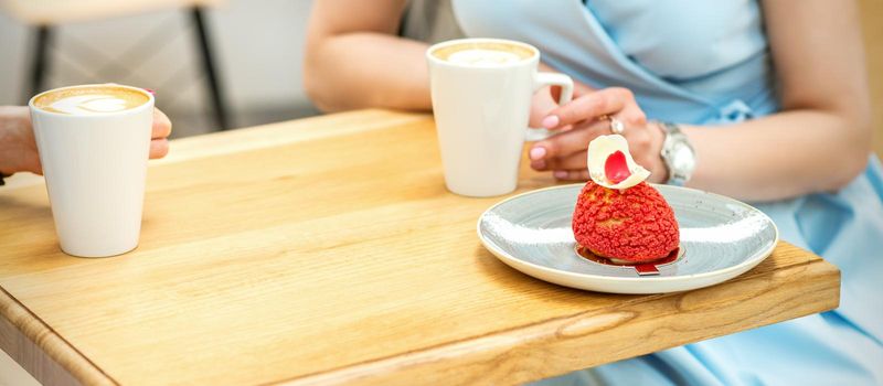 Two young girlfriends drinking coffee with pieces of cake sitting at the table in a cafe outdoors