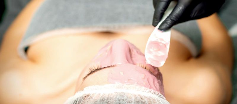 The cosmetologist applying an alginic mask to the face of a young woman in a beauty salon