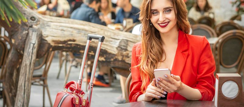 Beautiful young travel woman with smartphone sitting at the table looking at camera in cafe outdoors
