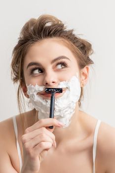 Beautiful young caucasian smiling woman shaving her face with razor looking up on white background