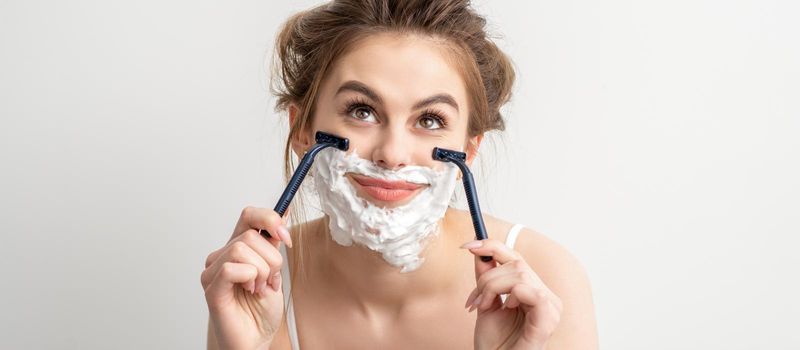 Beautiful young caucasian smiling woman shaving her face with razor looking up on white background