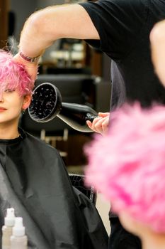 A male hairdresser professional drying stylish pink hair of the female client with a blow dryer in a beauty salon