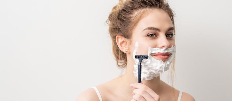 Beautiful young caucasian smiling woman shaving her face with razor on white background