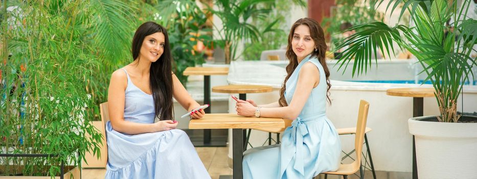 Two beautiful young women sitting at the table with smartphones looking at camera in a cafe outdoors