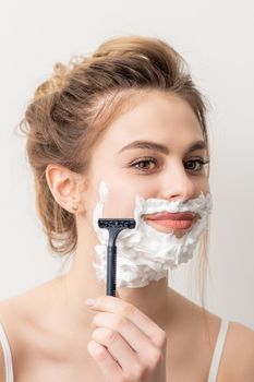 Beautiful young caucasian smiling woman shaving her face with razor on white background