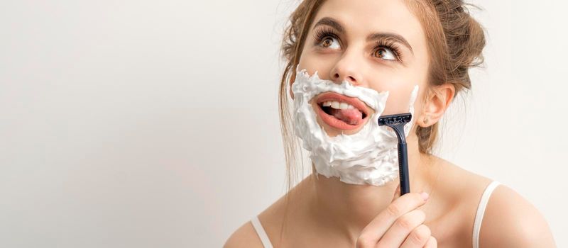 Beautiful young caucasian smiling woman shaving her face with razor sticking out tongue looking up on white background