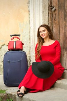 A young caucasian woman is sitting on the threshold of an old house in a red long dress with suitcase looking at the camera outdoors