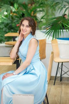 Beautiful smiling caucasian young woman in long blue dress sitting at the table and looking at the camera in a cafe outdoors