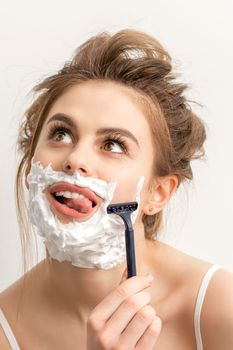Beautiful young caucasian smiling woman shaving her face with razor sticking out tongue looking up on white background
