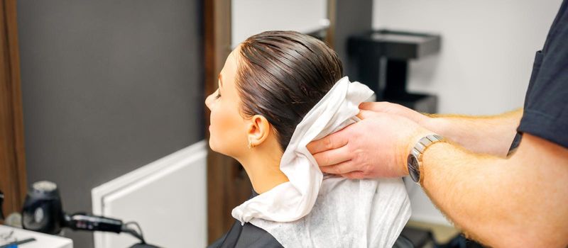 A male hairdresser's hands wipe the hair of a female client with a white towel after shampooing in a beauty salon