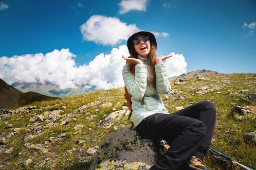 A smiling, positive brunette against a blue sky with fluffy white curly clouds sits on a stone in the mountains.