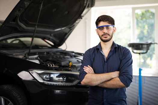 Portrait Bearded Handsome Car Mechanic is Posing in a Car Service. He Wears a Jeans Shirt and Safety Glasses. His Arms are Crossed. Specialist Manager Looks at a Camera and Smiles.