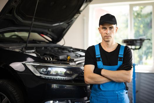 Portrait of a car mechanic crosses hands in a car workshop in blue uniform with equipment looking into camera.
