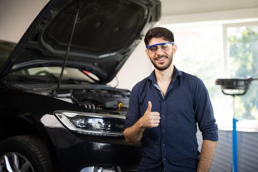 Happy technician bearded brunet mechanic man 20s in safety glasses work in vehicle repair shop show thumb up like gesture on light workshop background indoors.