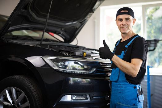 Positive auto service worker smiling to camera and showing thumb up gesture, approving car repair workshop. Portrait of car mechanic in a car workshop shows thumbs up, in the background of service.