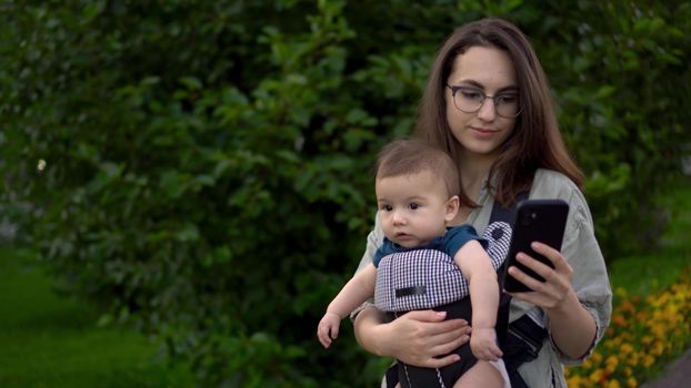 A young woman walks through the park with a child and a phone in her hands. Mom in glasses with her son in her arms. Newborn son in a kangaroo-backpack. 4k