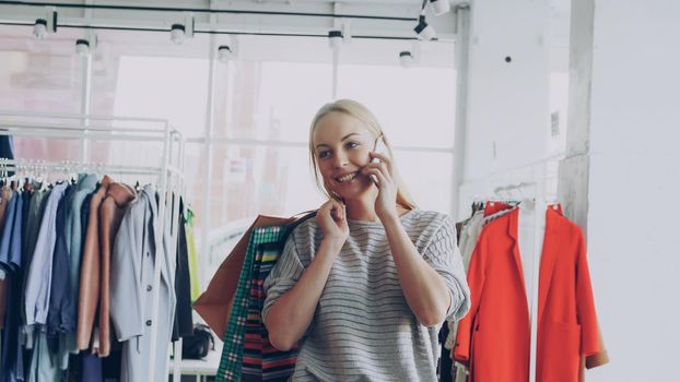Attractive blond young woman is walking between shelves and rails in large clothing store and talking on mobile phone. She is carrying bags, smiling and looking at trendy clothes around her.