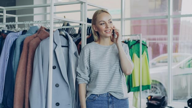 Cute blond girl is chatting on smart phone in modern boutique, touching clothes on hangers and smiling charmingly. Lots of fashionable clothes and large window in background.