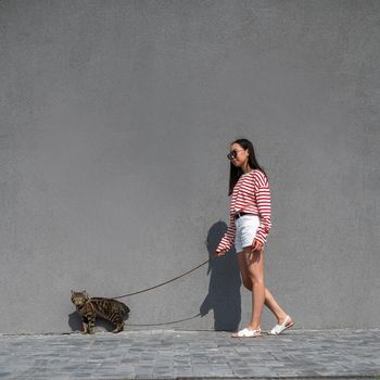 A young woman walks with a gray tabby cat on a leash against a gray wall