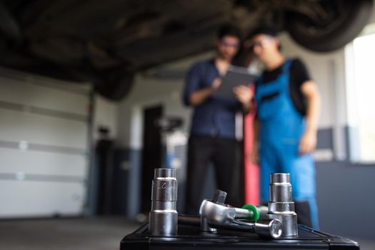 Male Mechanic Talking to Manager Under Vehicle in Car Service. Specialist is Showing Info on a Tablet Computer. Empowering Man in Modern Workshop.