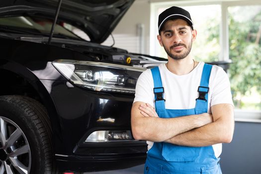 Young caucasian bearded man in blue overalls and black cap looks into camera, while smiling and crosses arms. Male car mechanic at workplace in spacious repair shop.