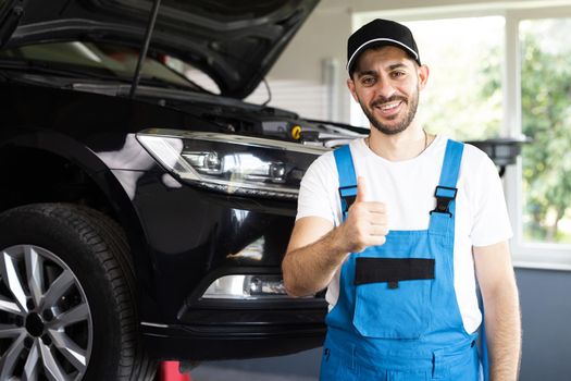 Portrait of bearded car mechanic in a car workshop shows thumbs up. Positive auto service worker in blue overalls and cap smiling to camera and showing thumb up gesture, approving car repair workshop.