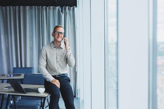 Young smiling businessman holding a cup of coffee in his hands in the office businessman looking out the window standing in a modern workplace man taking a vacation from hard work