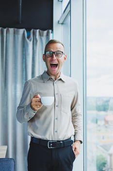 Modern office businessman or manager drinking coffee by the window, smiling. Portrait of successful IT engineer programmer wearing glasses working in office.