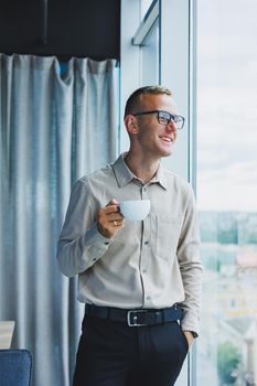 Modern office businessman or manager drinking coffee by the window, smiling. Portrait of successful IT engineer programmer wearing glasses working in office.