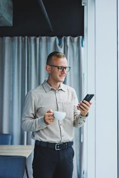 Young smiling businessman holding a cup of coffee in his hands in the office businessman looking out the window standing in a modern workplace man taking a vacation from hard work