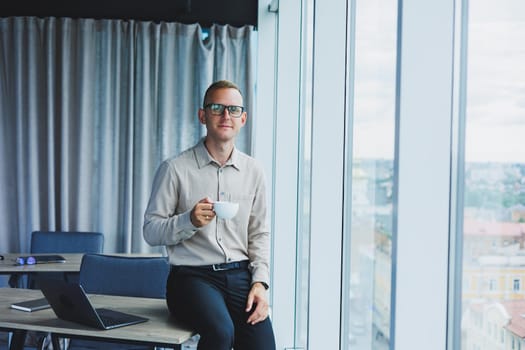 Young smiling businessman holding a cup of coffee in his hands in the office businessman looking out the window standing in a modern workplace man taking a vacation from hard work