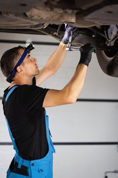 Male Mechanic Working on Vehicle in Car Service. Empowering Man Wearing Gloves and Using Ratchet Underneath the Car. Modern Workshop.