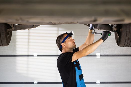 Young caucasian man in gloves and blue coveralls tightens bolt under car using ratchet. Male car mechanic at work in spacious repair shop. Modern workshop.