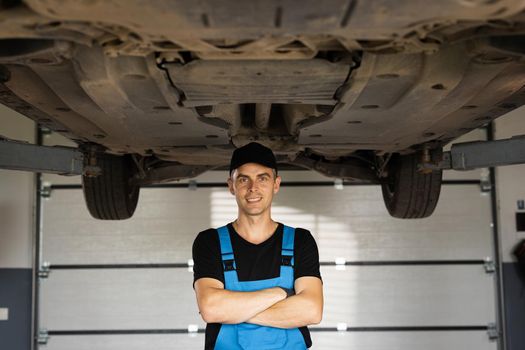 Portrait of mechanic repairing in uniform standing looking camera crossed hands at under lifted car of automobile. Repairing car service concept.