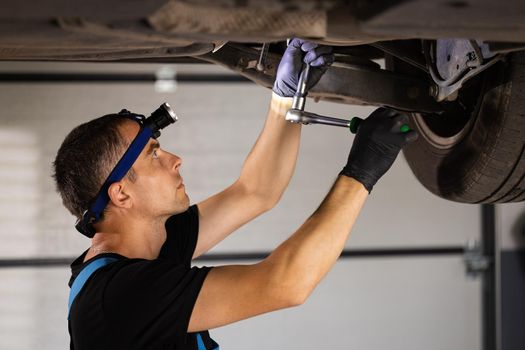 Professional car mechanic screwing details of car with special tool on lifted automobile at repair service station. Skillful man in uniform fixing car. Car service, repair maintenance.