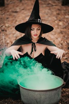 Black witch sits in front of cauldron from which smoke is falling. Sorceress conjures, brews potion with spells. Horror, halloween, cosplay holiday, magic concept. High quality photo