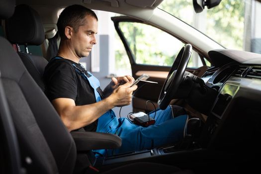 Mechanic sitting in car and working on laptop while doing vehicle diagnostic test in auto service garage. Automotive mechanic running diagnostics software on tablet computer