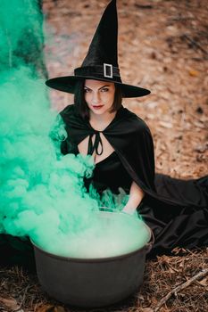 Black witch sits in front of cauldron from which smoke is falling. Sorceress conjures, brews potion with spells. Horror, halloween, cosplay holiday, magic concept. High quality photo