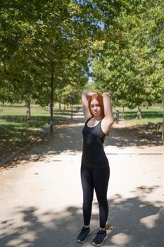 Full body fit young sportswoman stretching shoulder and looking at camera while warming up during fitness workout on sunny summer day in park