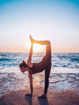 Slim woman in black bodysuit practicing yoga near sea or ocean during sunrise light. Flexibility, stretching, fitness, healthy lifestyle. High quality photo