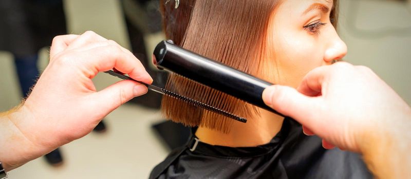 Hair stylist's hands straightening short hair of young brunette woman with flat iron and comb in a beauty salon