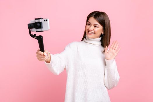 happy woman blogger using smart phone and steadicam for streaming, waving hand, greeting followers, wearing white casual style sweater. Indoor studio shot isolated on pink background.