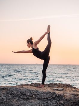 Slim woman in black bodysuit practicing yoga near sea or ocean during sunrise light. Flexibility, stretching, fitness, healthy lifestyle. High quality photo