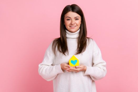 Beautiful woman has dream and holding paper small house, planning new home to buy, looks at camera, wearing white casual style sweater. Indoor studio shot isolated on pink background.